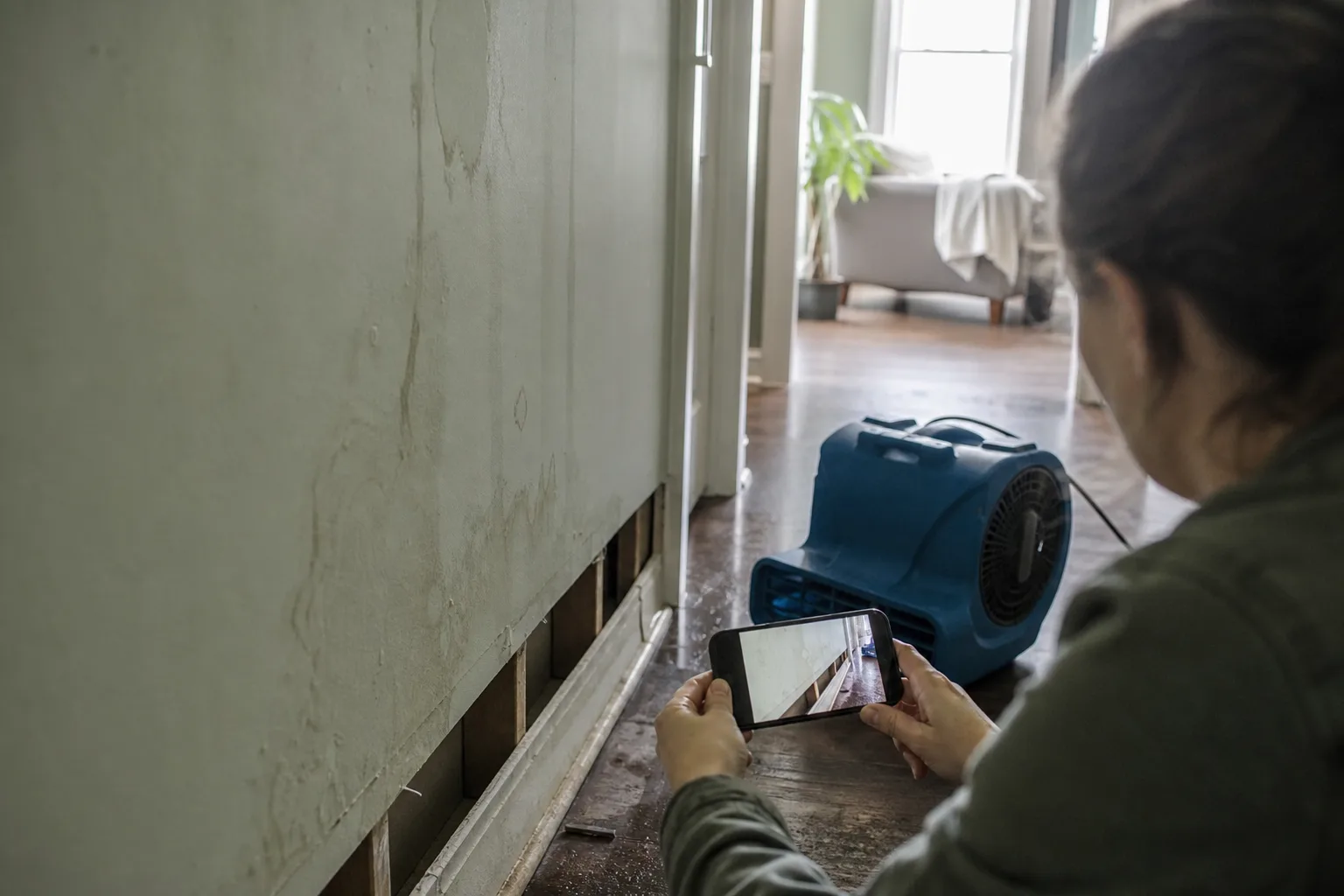 A homeowner photographing a water-stained wall and lifted baseboard with a smartphone while an air mover runs on the floor of a residential hallway