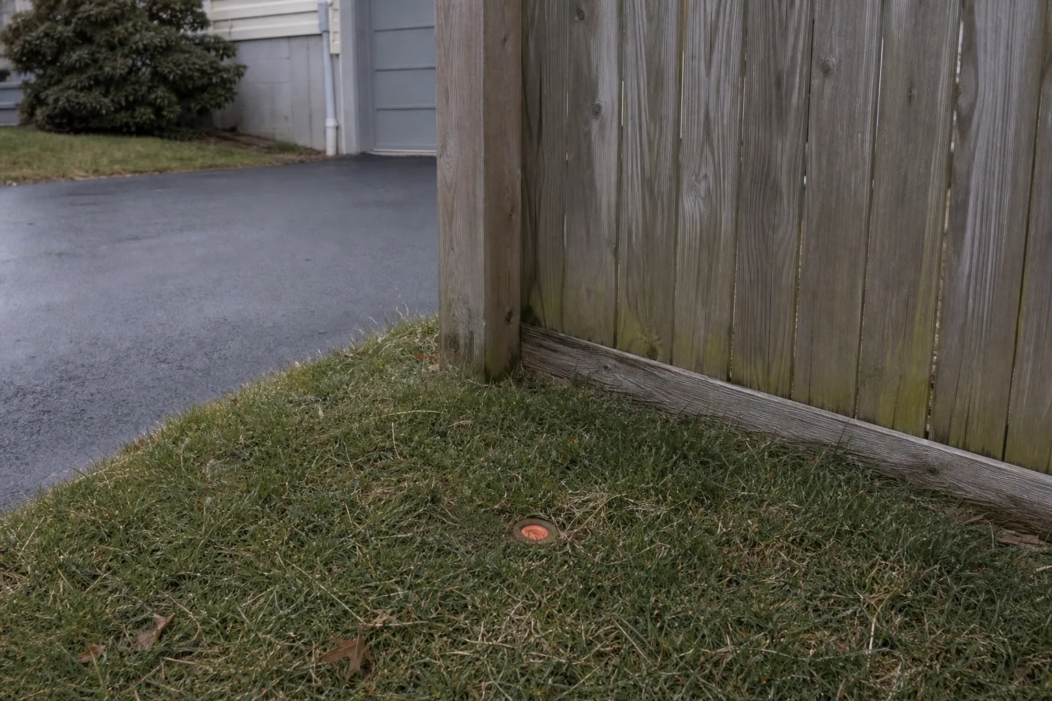 A surveyor's iron pin with an orange plastic cap set flush in grass at the corner of a residential lot, wooden fence and neighboring driveway visible behind it